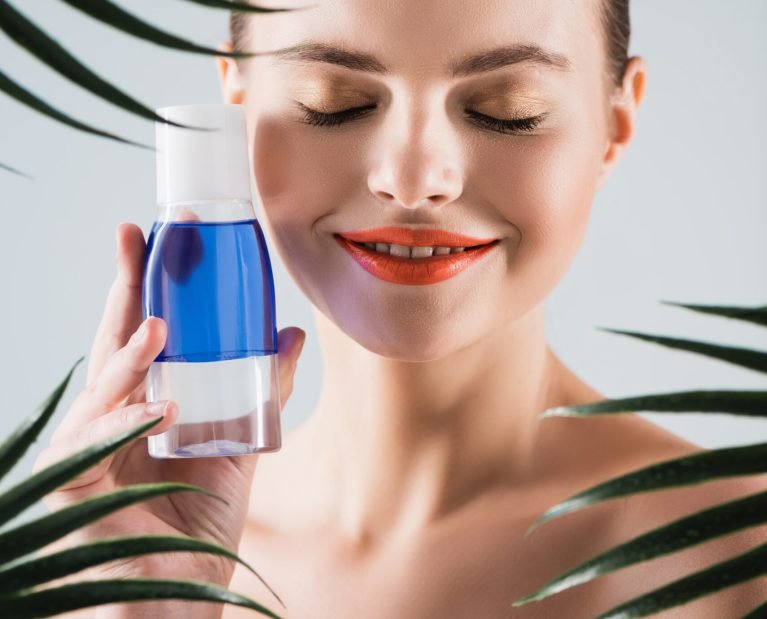 happy woman with makeup holding bottle with toner near palm leaves on white
